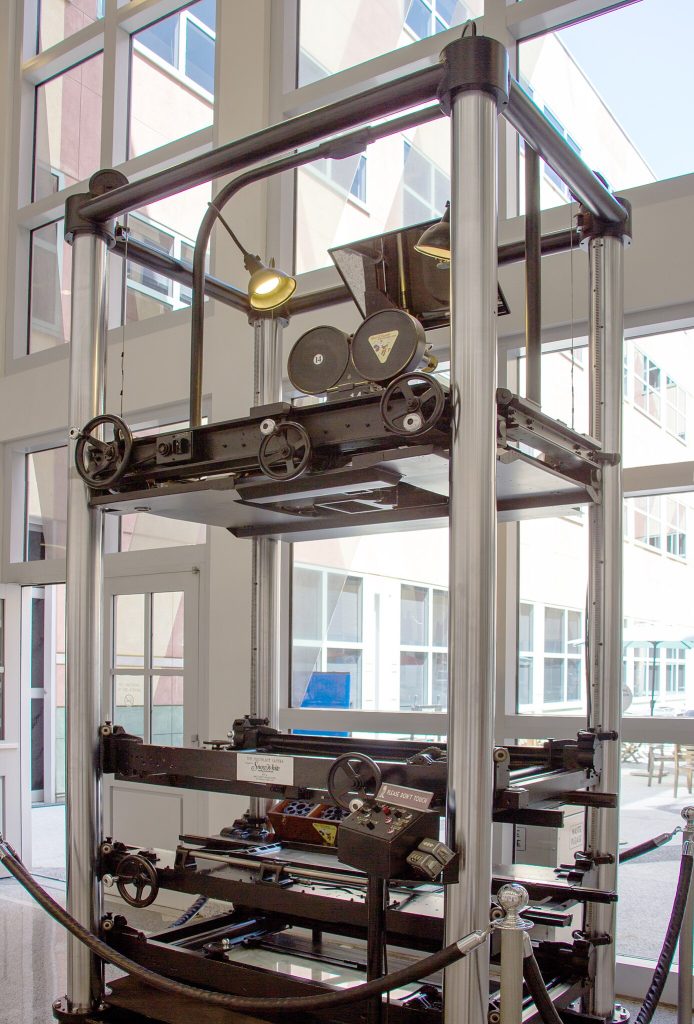 Machine consisting of multiple trays on mechanical rollers, holding planes of glass, with a camera on top.