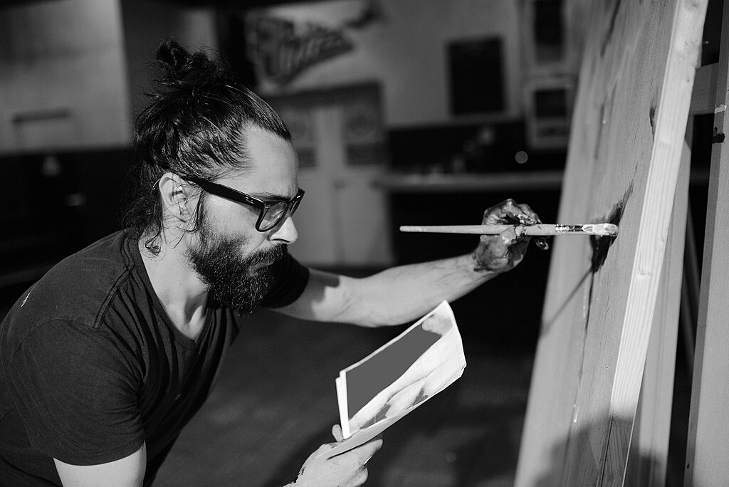 Man brush-painting on a canvas, with a paper sketch in hand.