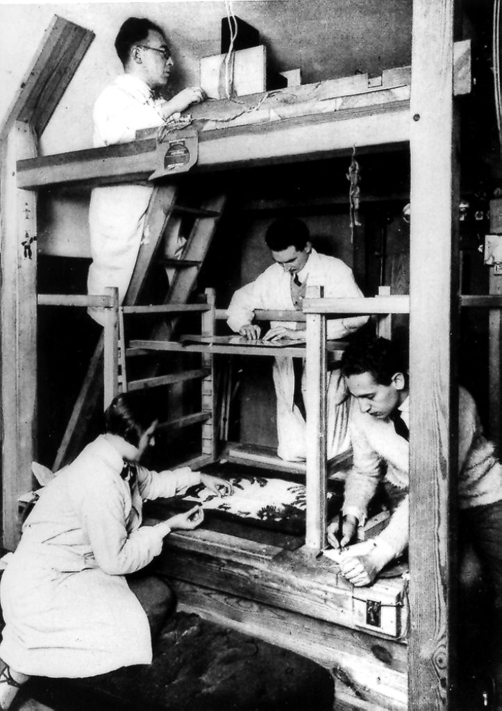 A wooden structure with glass planes, background, and camera, with three people working placing art on it.